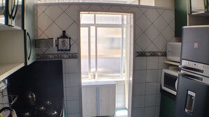 A compact kitchen with a sink, stove, and microwave. There is a window with blinds and a door leading to an outdoor balcony. - Between the beaches (Florianópolis)