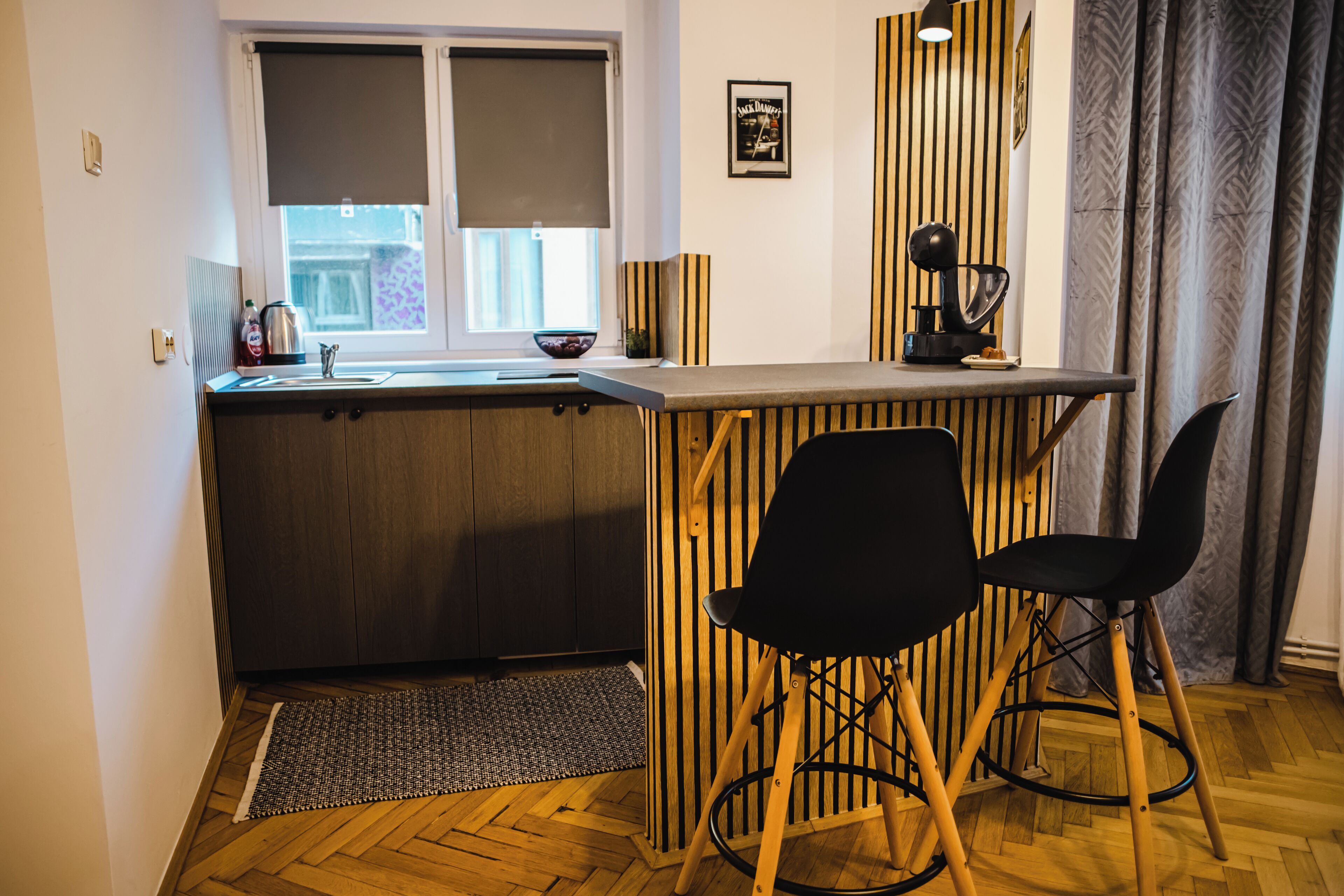 Una cocina compacta con mostrador de madera, dos sillas negras, un lavamanos pequeño y una ventana con persianas.