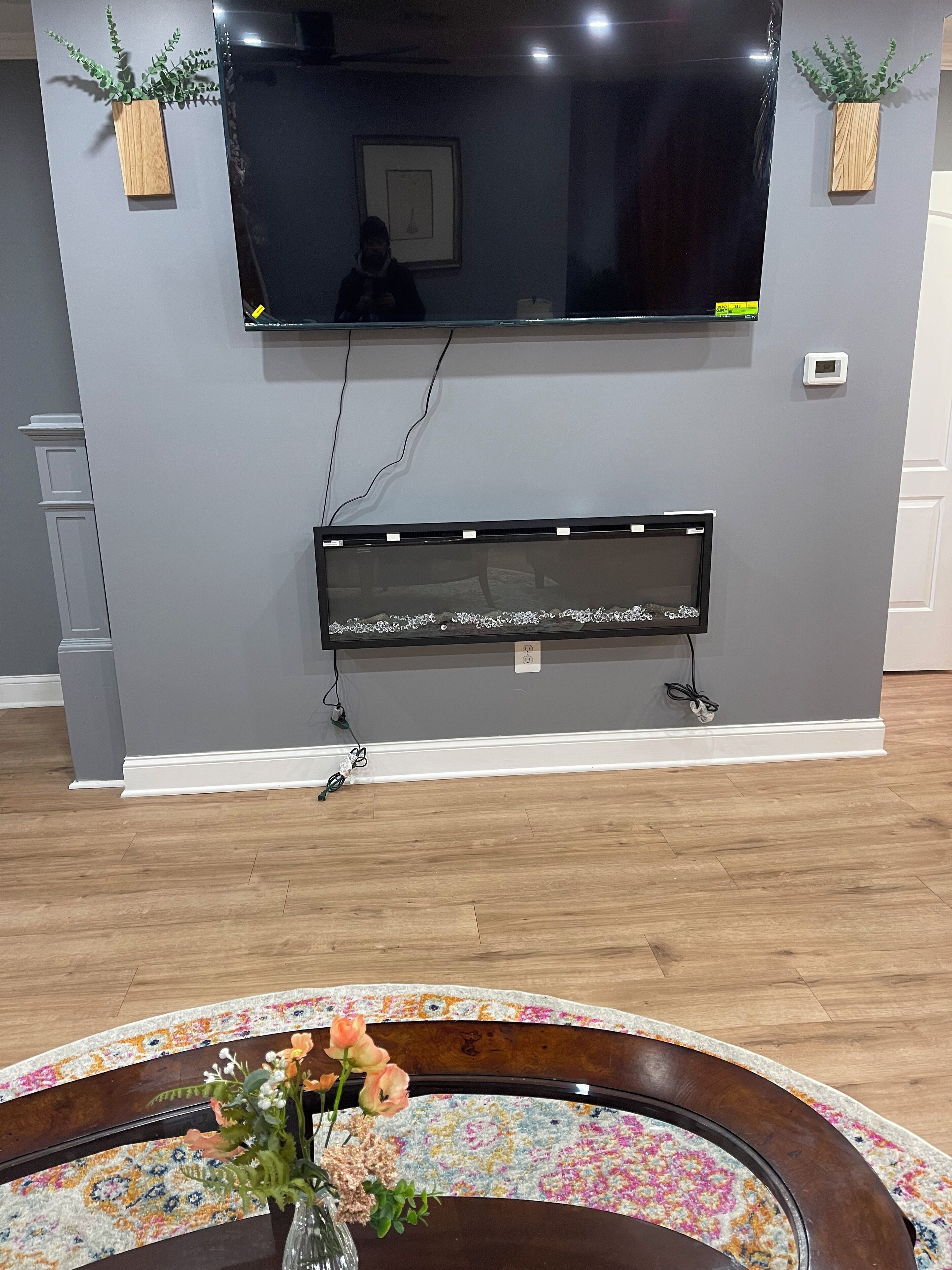 A living room with a flat-screen TV mounted on the wall, a fireplace below, and a floral arrangement on a round table.