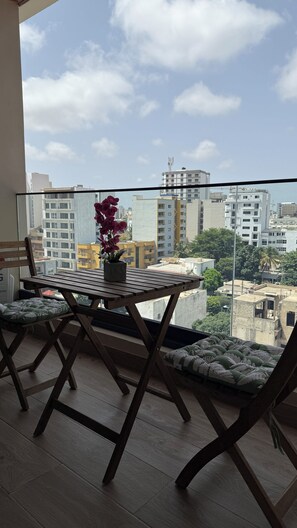 A balcony with a wooden table and chairs, a potted plant, and a view of urban buildings.