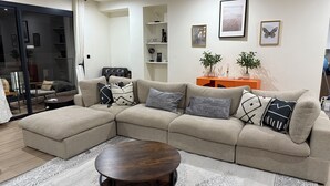 A modern living room with a sectional sofa, a round coffee table, and decorative pillows. There are framed pictures on the wall, a sideboard with plants, and a glass door leading to another room. - Elegant 3-bed, 3-bath, located in the desirable neighborhood of Point E, Dakar (Dakar)