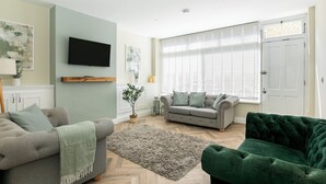 A living room with a grey sofa, a green armchair, a grey sofa, a wooden TV stand, a grey rug, and a large window with blinds. - Historic Barber Shop, Parking, Garden, Town Centre (Cirencester)