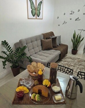 Una sala de estar con un sofá gris, una mesa de comedor con una bandeja de frutas, una cafetera y una planta en maceta.