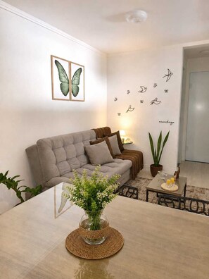 Una sala de estar con un sofá gris, una mesa de centro con un jarrón de flores, una pared decorada con adhesivos de mariposas y pájaros, y una planta en maceta.