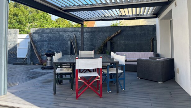 Eine überdachte Terrasse mit Tisch, Stühlen, einem Grill und einer Sitzecke.