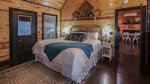 Un dormitorio acogedor con techo de madera, una cama con manta azul, una mesita de noche con lámpara y un estante en la pared con objetos decorativos.
