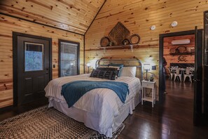 A cozy bedroom with a wooden ceiling, a bed with a blue blanket, a bedside table with a lamp, and a wall-mounted shelf with decorative items. - Welcome to Goodnight Moon (Broken Bow)
