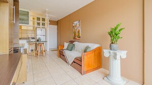 Un salon avec un canapé, une table basse et une plante en pot. Il y a également un coin cuisine avec des placards et un plan de travail.