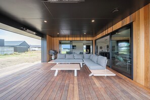 A modern outdoor living area with a wooden deck, a grey sectional sofa, and a white table. - Breaksea Basecamp - Te Anau Holiday Home (Te Anau)