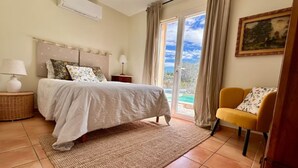 Ein Schlafzimmer mit Bett, Nachttischen, einem Sessel, einem Gemälde und Blick durch eine Glastür.