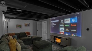 A modern living room with a large flat-screen TV displaying a Roku TV interface, a fireplace, and a sectional sofa with multiple cushions.