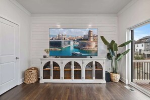 Una habitación con un televisor de pantalla plana grande montado en una pared blanca, un mueble blanco con puertas arqueadas debajo y una planta en maceta a la derecha.