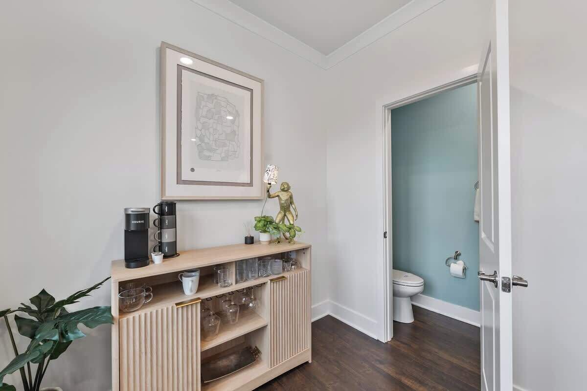 A hallway with a wooden console table holding a coffee maker, glasses, and a framed map. A door leads to a bathroom with a toilet and a towel rack.