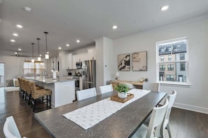 Una cocina moderna con armarios blancos, electrodomésticos de acero inoxidable y un comedor con una mesa larga y sillas.