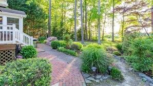 A garden with a brick pathway, a wooden deck, and various plants. - The Sandy Hook Retreat| Grill & Sunroom (Ocean Pines)