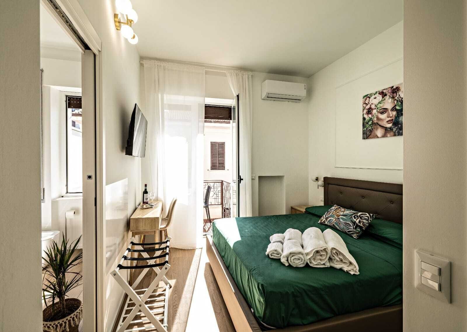A hotel room with a bed, a desk, a chair, a potted plant, and a painting on the wall.