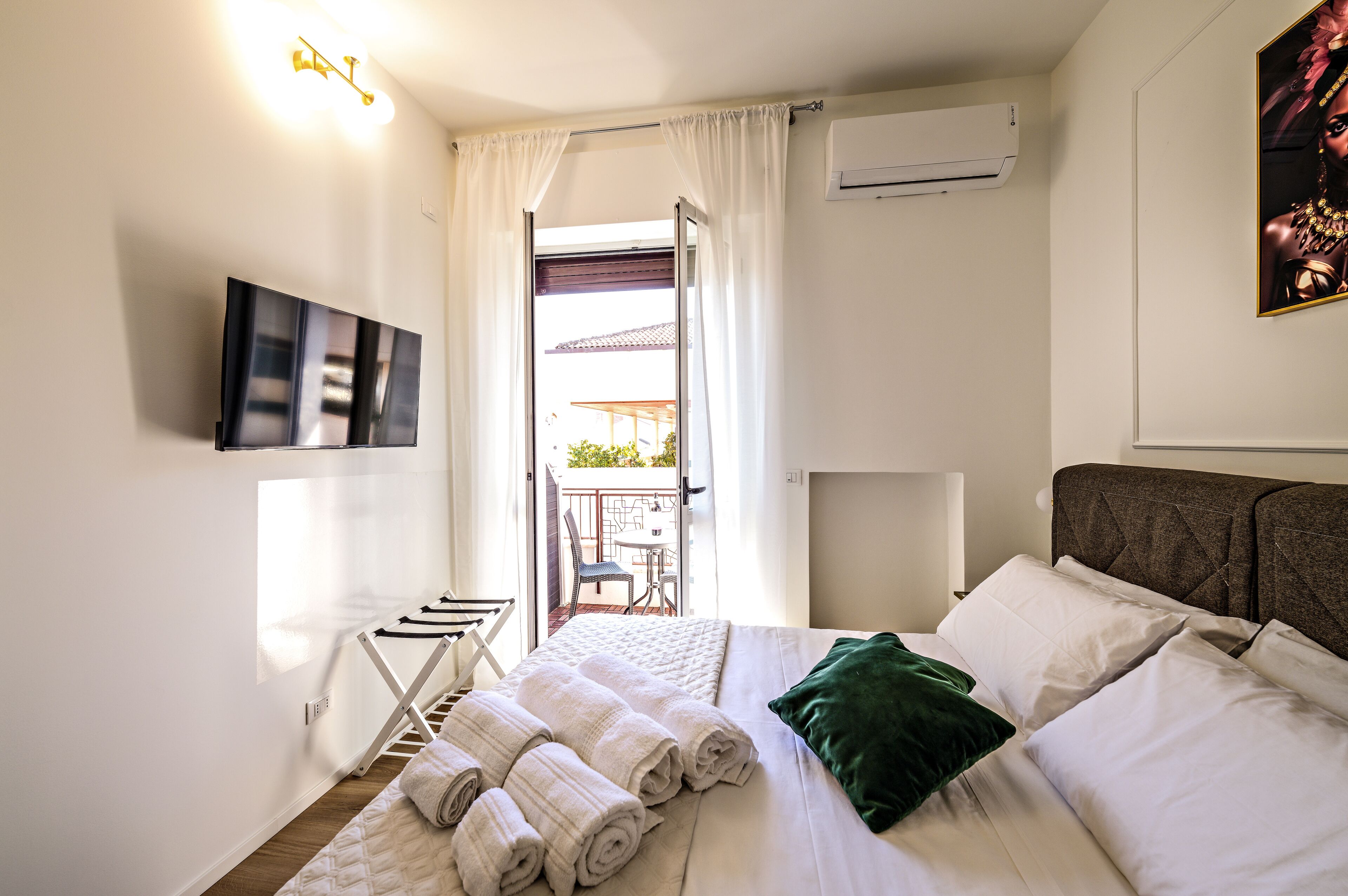 A hotel room with a bed, a TV mounted on the wall, a balcony with a table and chairs, and an air conditioning unit.