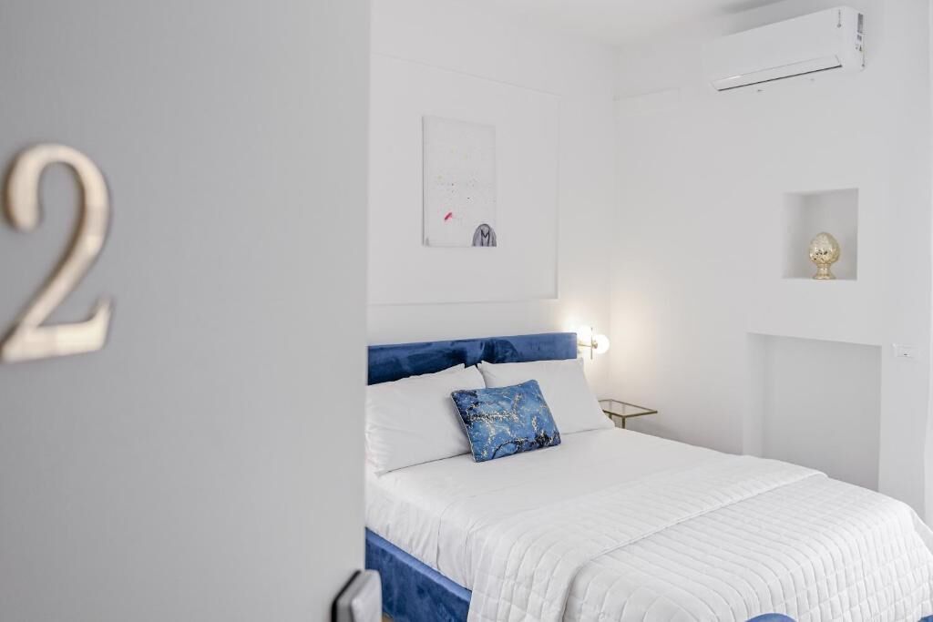 A bedroom with a bed, a wall-mounted air conditioner, a small shelf with a decorative item, and a framed picture on the wall.