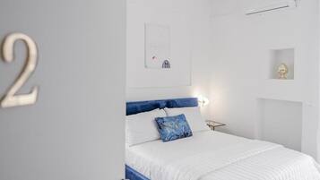 A bedroom with a bed, a wall-mounted air conditioner, a small shelf with a decorative item, and a framed picture on the wall.