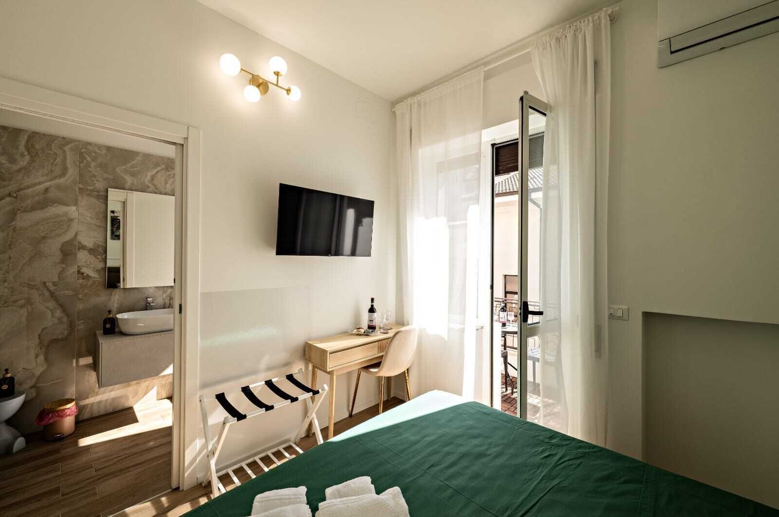 A modern bedroom with a green bed, a wooden desk, a chair, a TV, and a bathroom with a marble wall and a sink.
