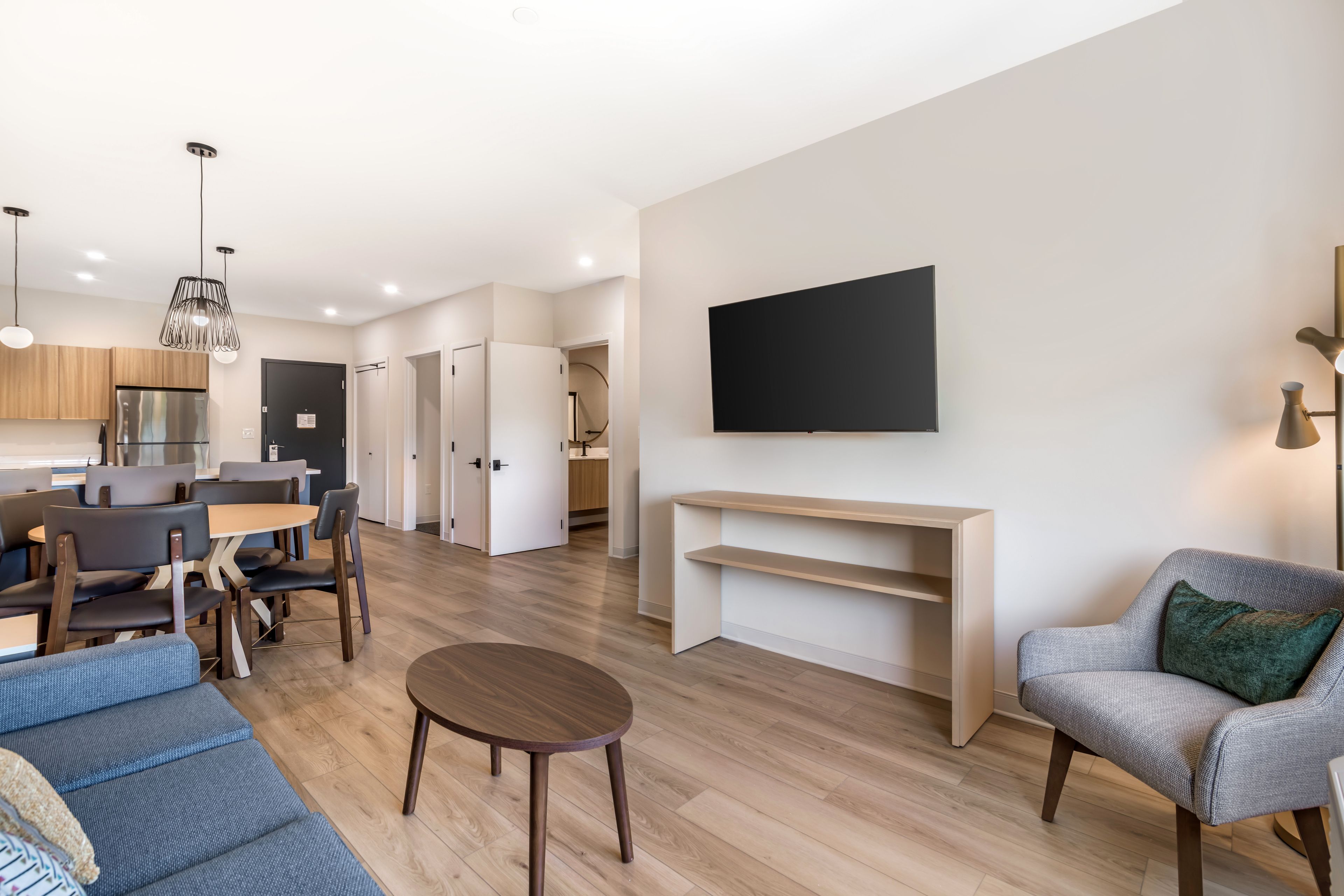 A modern living room with a sofa, a coffee table, a TV mounted on the wall, and a dining area with chairs and a table.