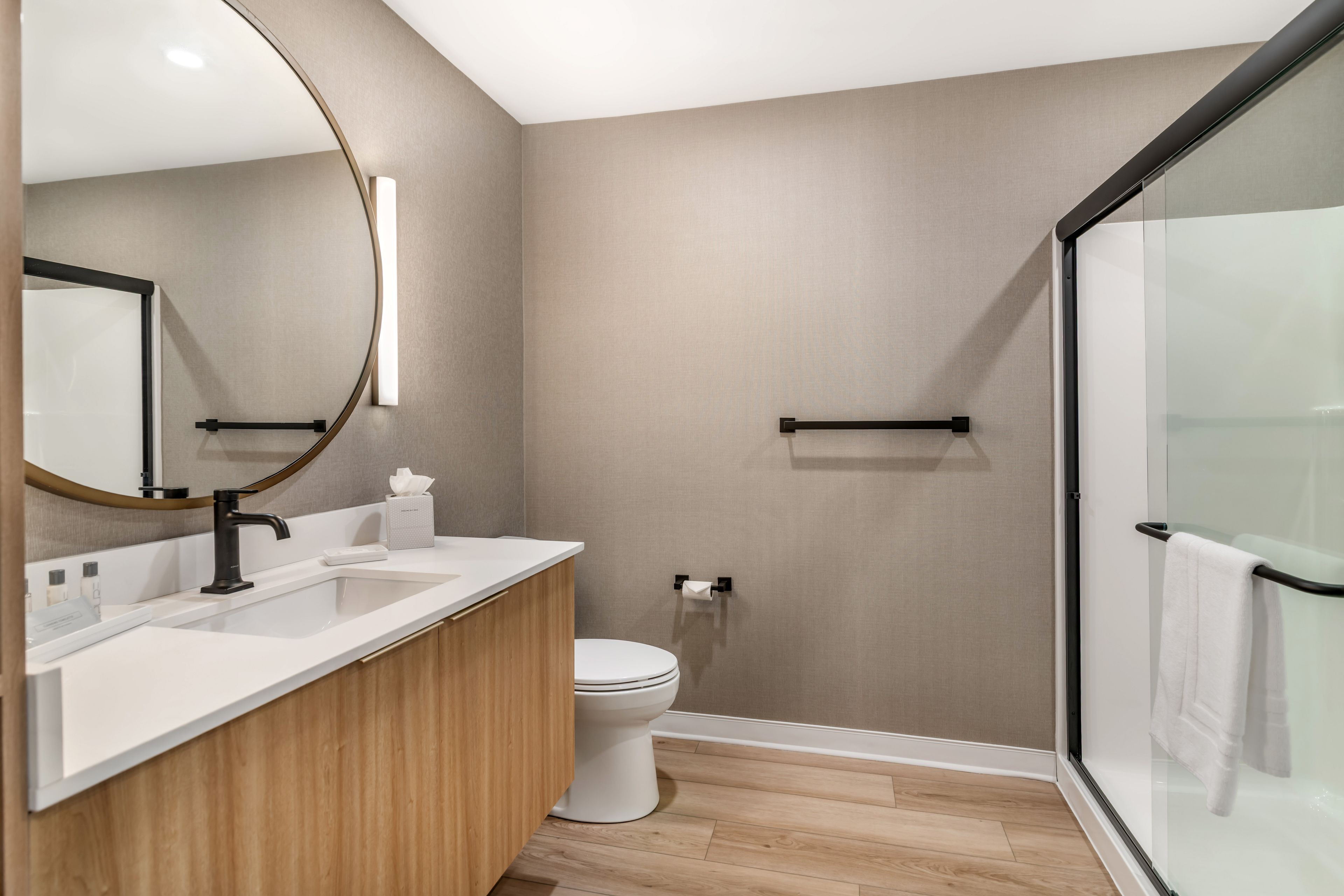 Ein modernes Badezimmer mit einem Holz-Waschtisch, einem runden Spiegel, einer Toilette und einer Duschkabine aus Glas.