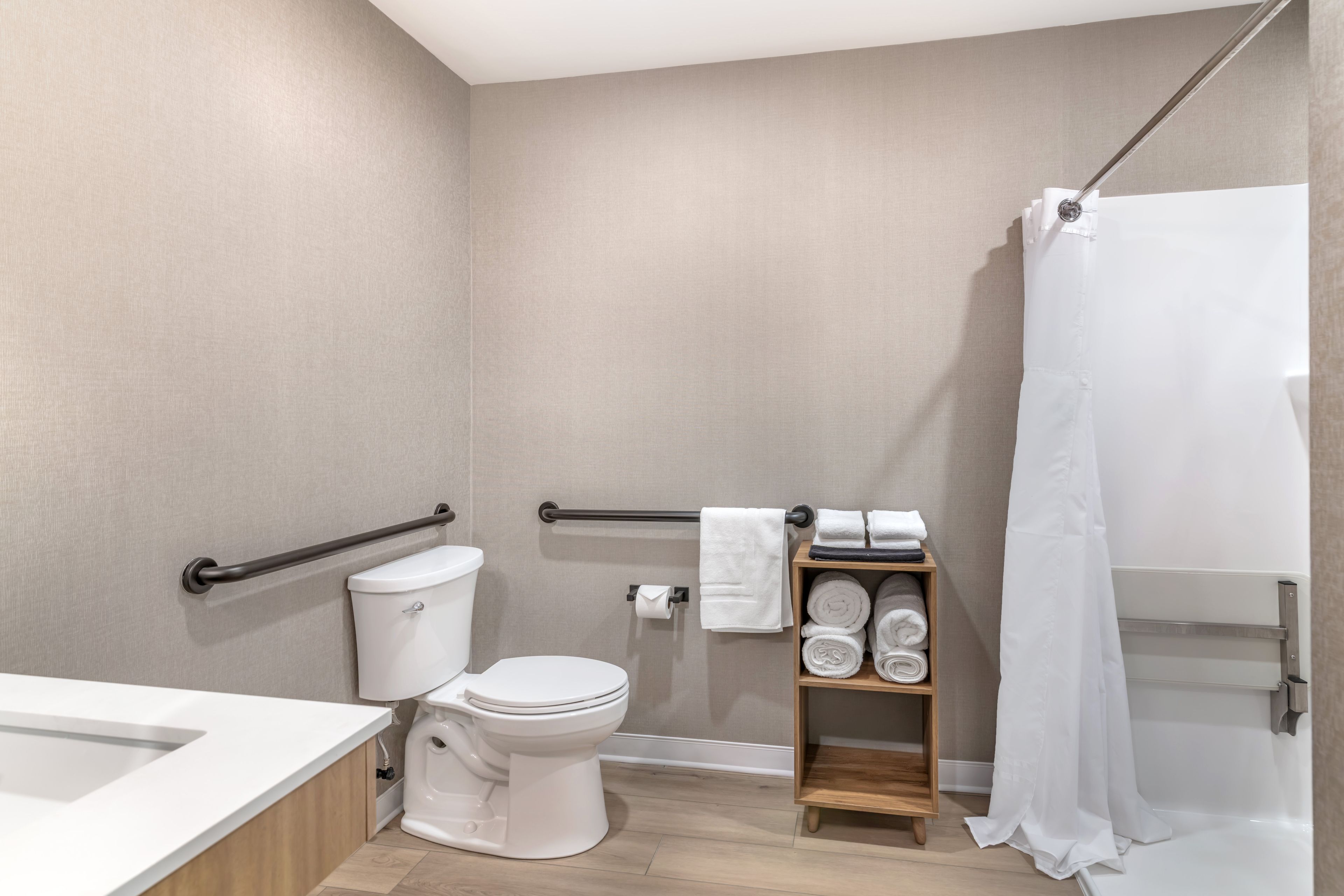Un baño moderno con inodoro, ducha con cortina, estante de madera con toallas y barra de apoyo.