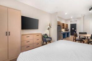 A modern hotel room with a large bed, a flat-screen TV mounted on the wall, a desk with a chair, and a kitchenette area. - XStay Suites Mansfield (Mansfield)