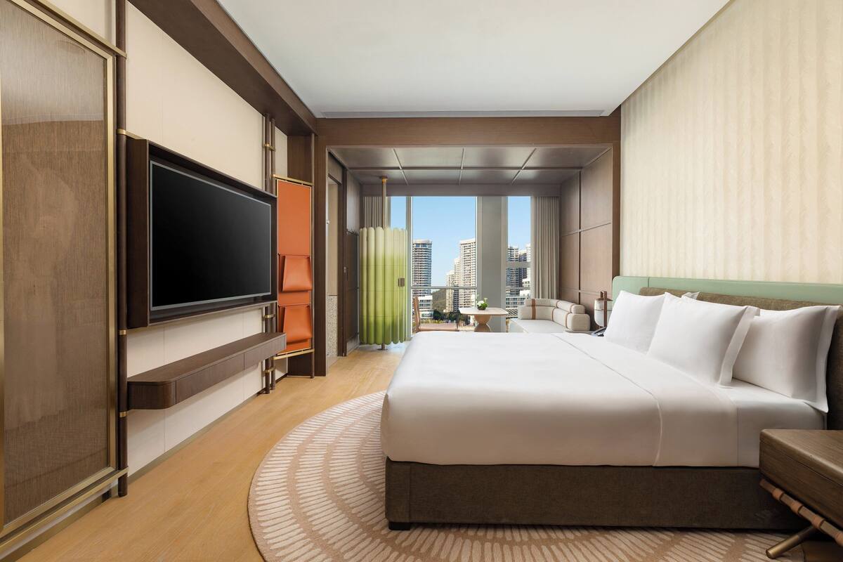 A modern hotel room with a large bed, a flat-screen TV, a city view, and a small table with a plant.