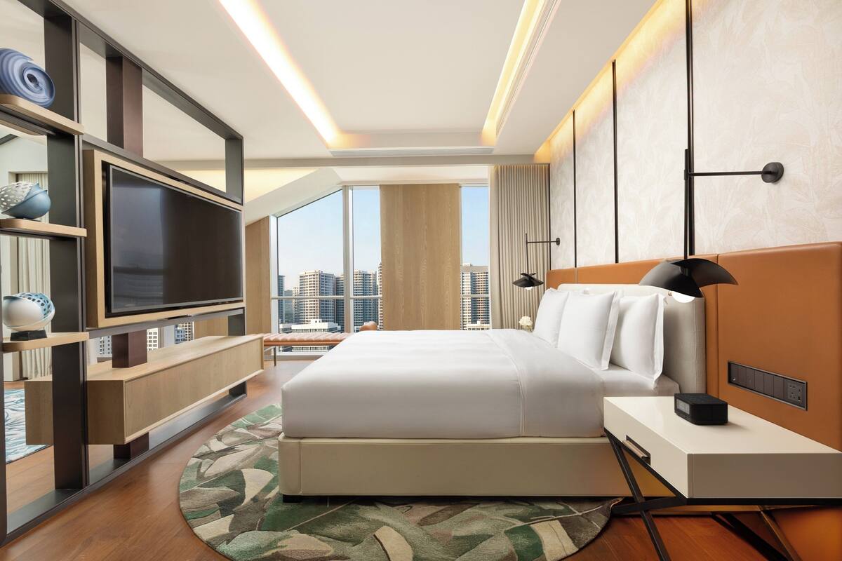 A modern hotel room with a large bed, a flat-screen TV, a bedside table, a bookshelf, and a city view.