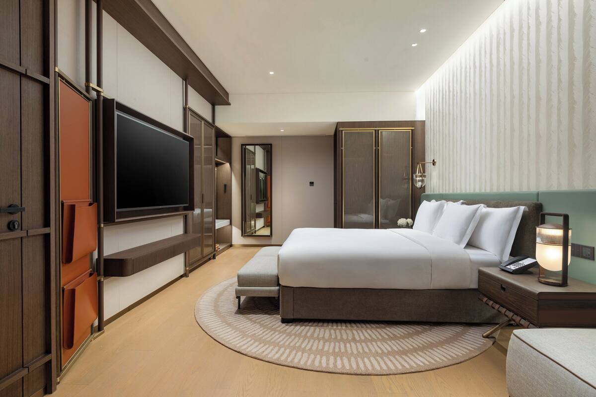 A modern hotel room with a large bed, a flat-screen TV, a wardrobe, and a bedside table with a lamp.