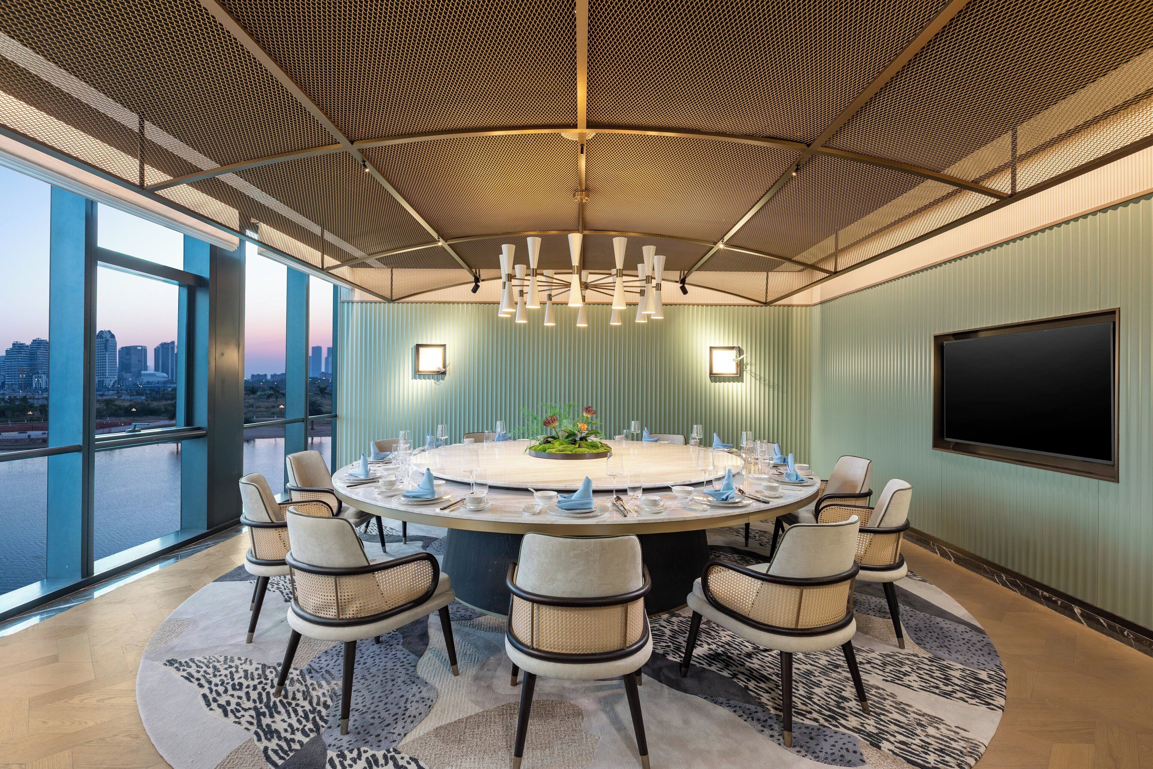 A conference room with a round table, chairs, a chandelier, a large window with a city view, and a flat-screen TV.