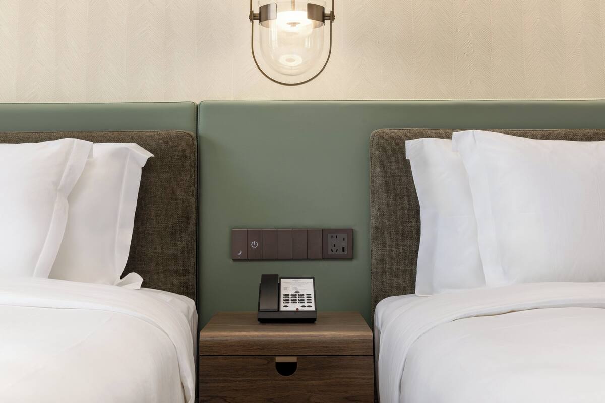A hotel room with two beds, a nightstand with a phone, and a wall-mounted light fixture.