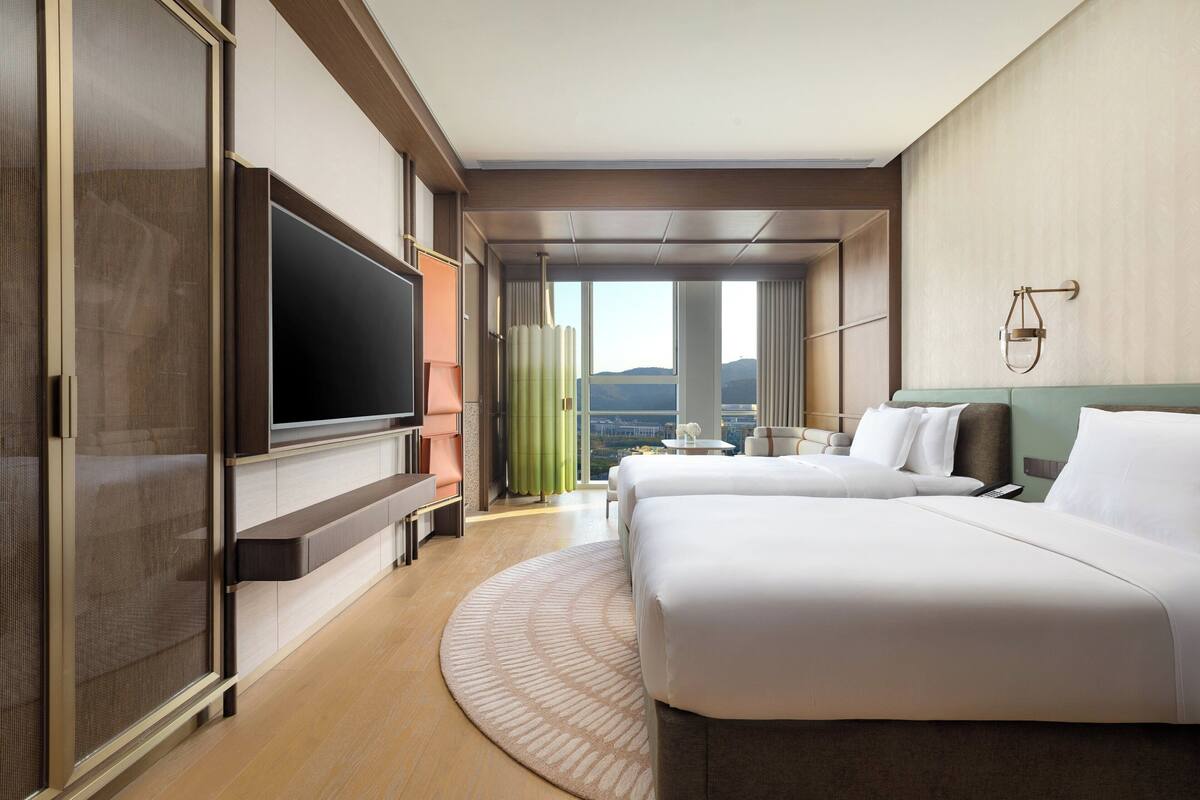 A hotel room with a large flat-screen TV, two beds, a round rug, and a view of the cityscape through large windows.