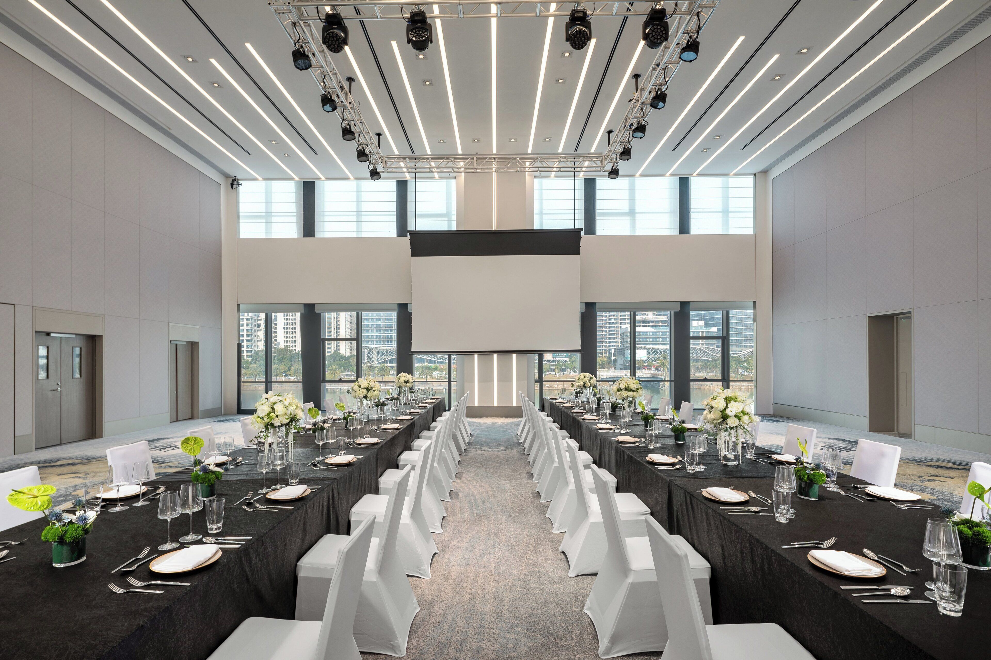 Un salón de conferencias con una larga mesa dispuesta para un evento formal, ventanas amplias y un techo moderno con luminarias.