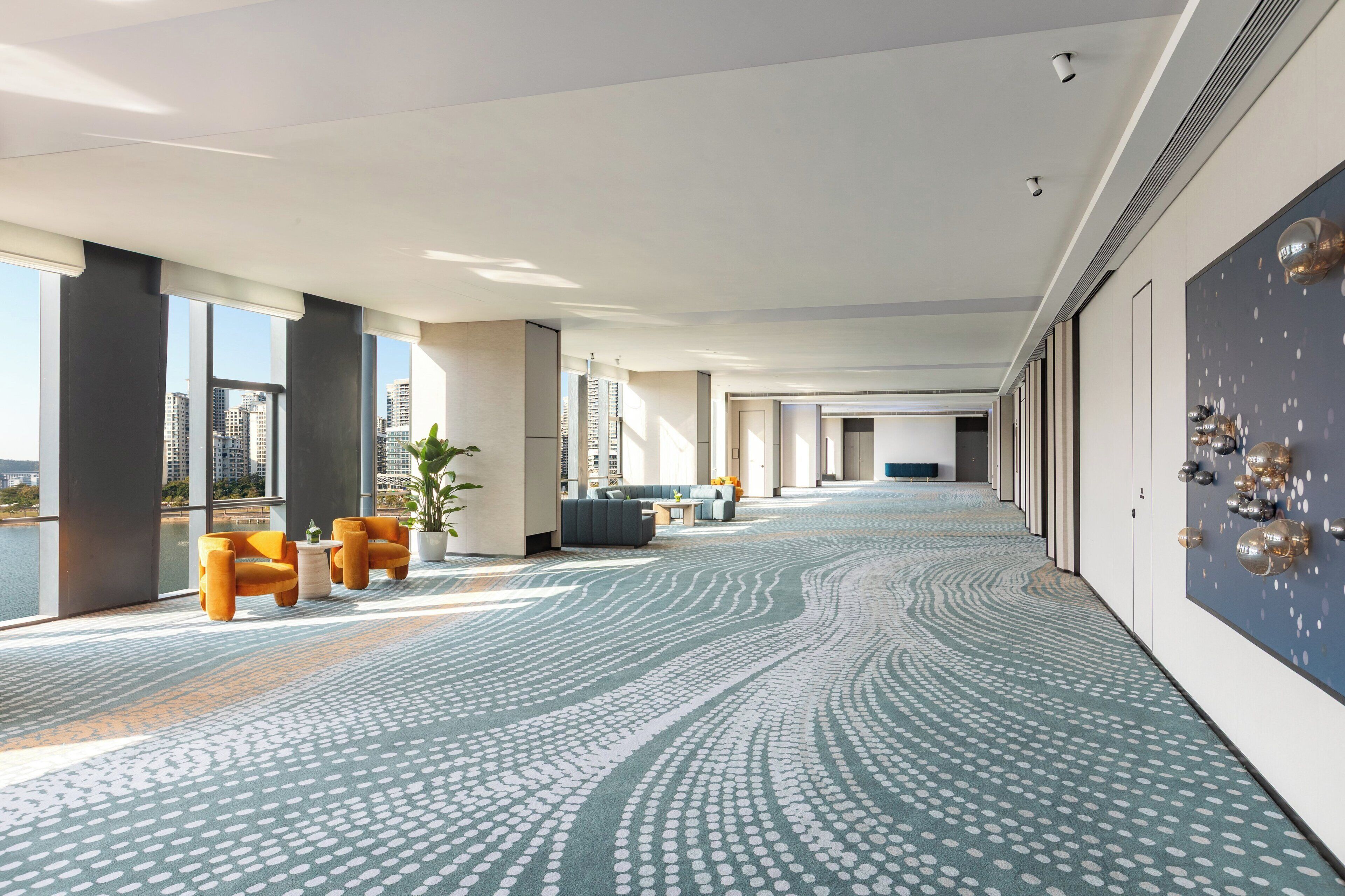 A spacious hotel corridor with a patterned carpet, large windows offering a city view, and modern furnishings.