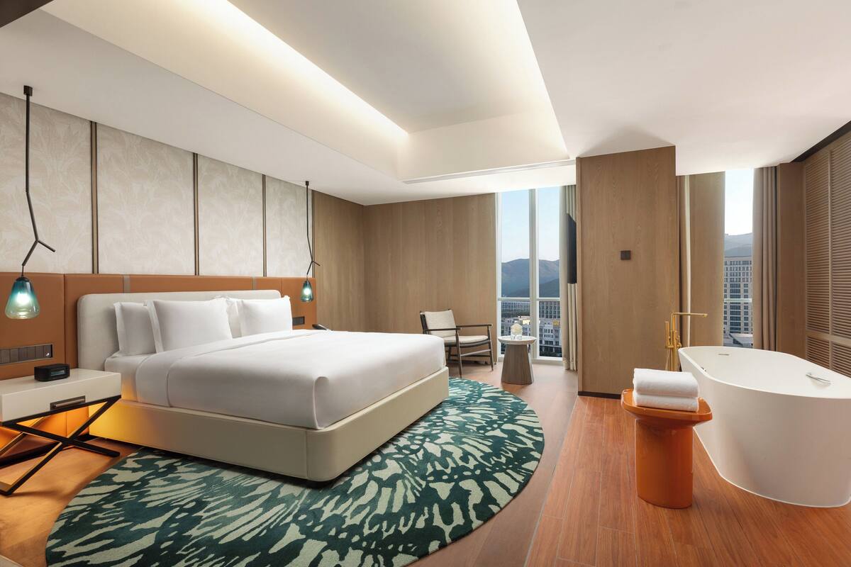 A modern hotel room with a large bed, a sitting area, a bathtub, and a city view.