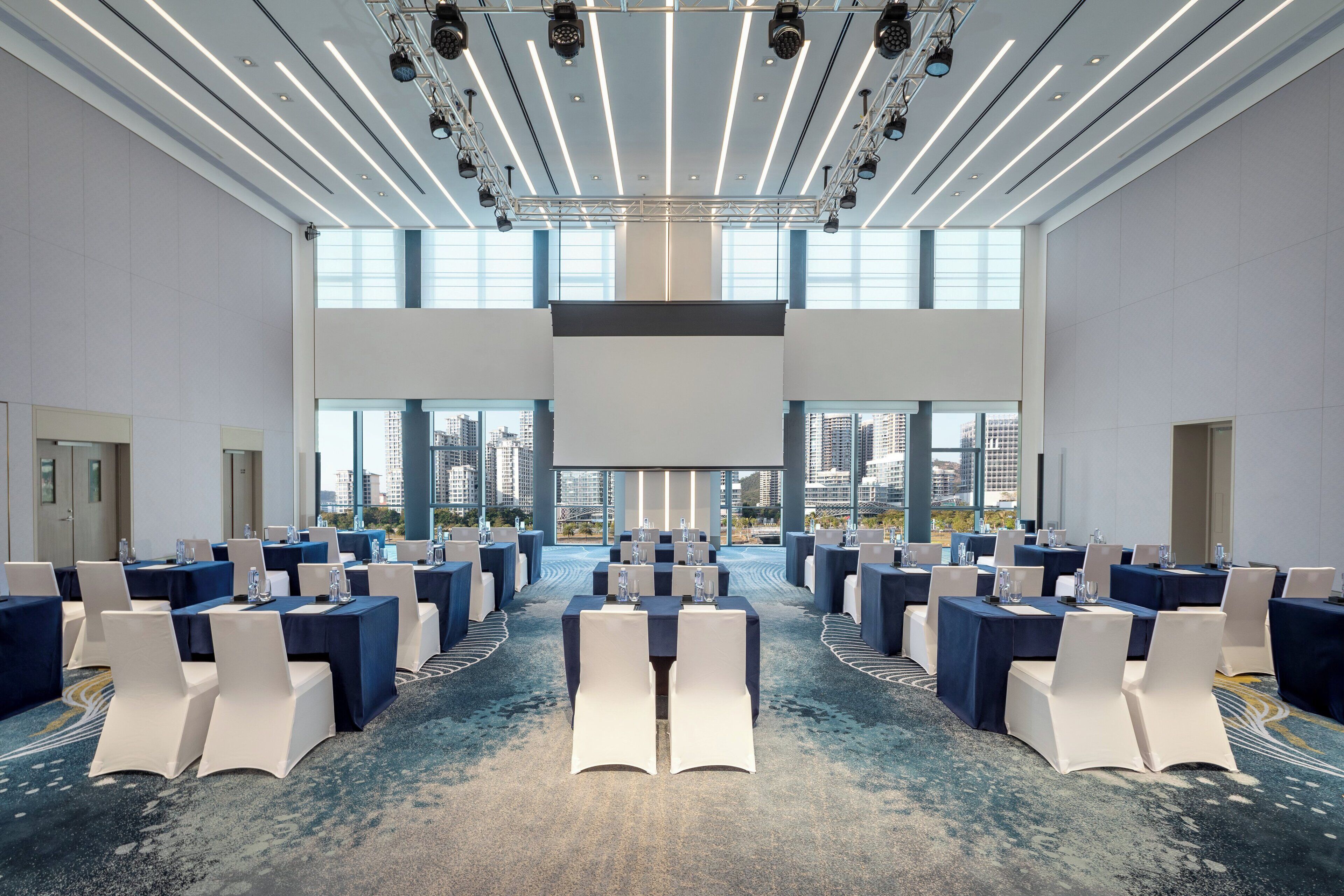 A large conference room with tables and chairs, high ceilings, and large windows offering city views.