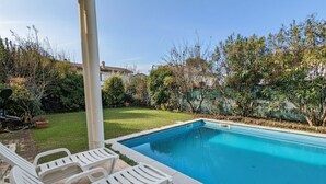 Una piscina con sillas reclinables blancas, rodeada de vegetación y un cielo despejado.