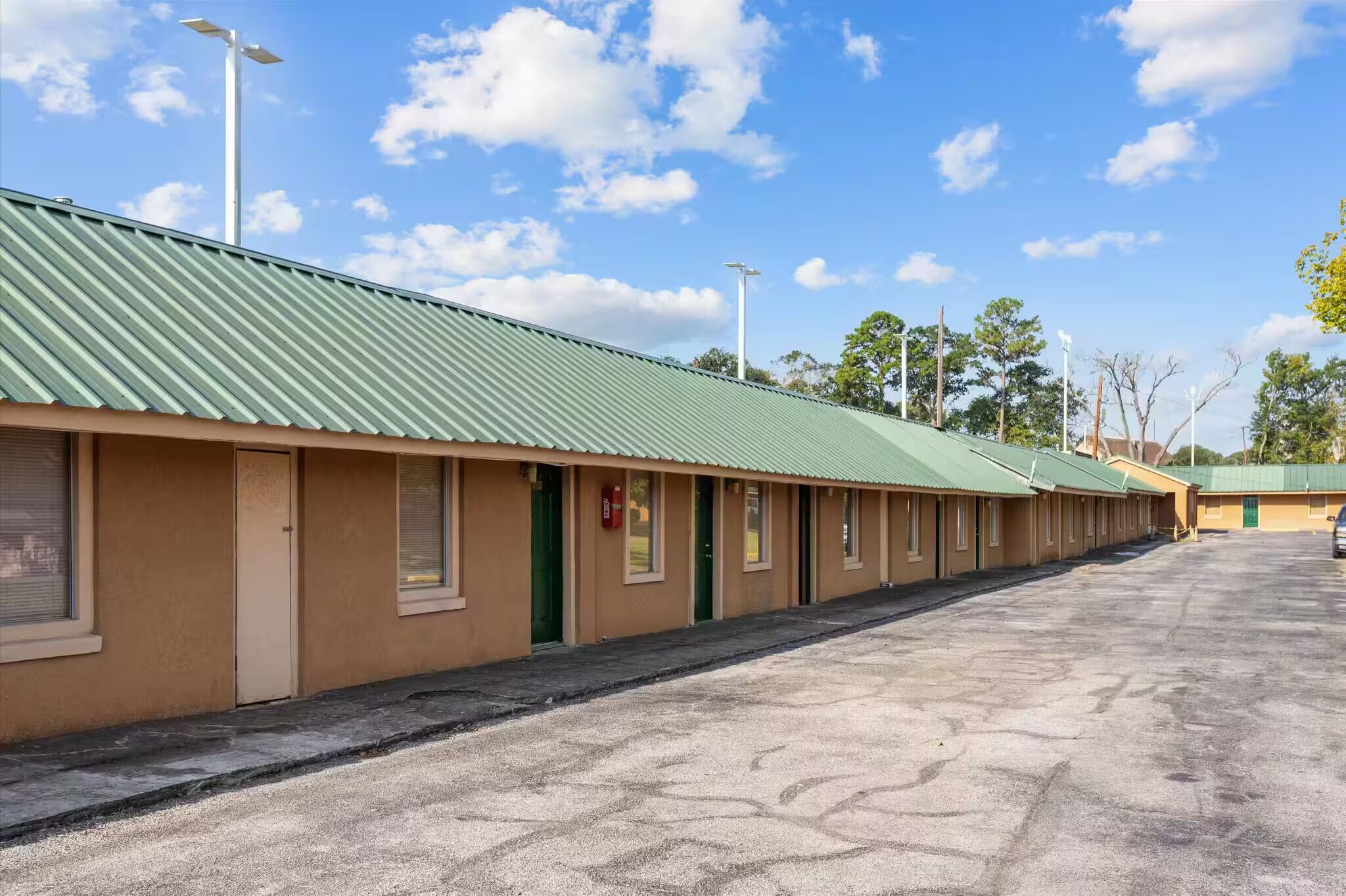 Une rangée de chambres de motel avec des toits verts et des murs de couleur beige, un parking et des arbres en arrière-plan.