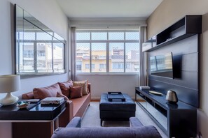 Un salon moderne avec un canapé, une table basse, un téléviseur et une grande fenêtre donnant sur la ville.