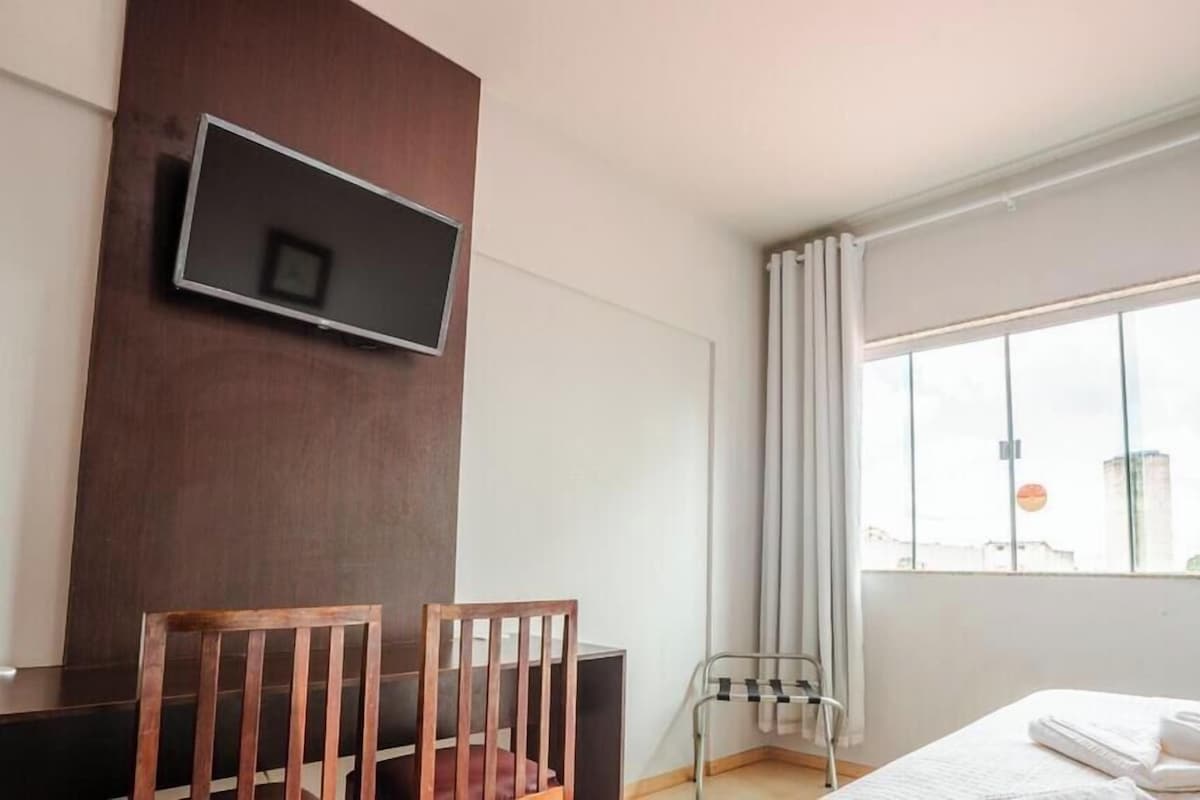 A hotel room with a bed, a TV mounted on the wall, a window with curtains, and a chair.