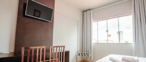 Habitación de hotel con una cama, televisión montada en la pared, ventana con cortinas y una silla.