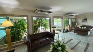 A spacious lobby with a brown leather sofa, a coffee table with a floral arrangement, and a large window offering a view of greenery. - Rede Andrade Aquarius (Porto Velho)