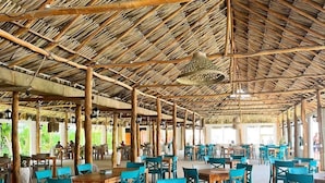 Un restaurante al aire libre con mesas y sillas de madera, techo de paja y luz natural.