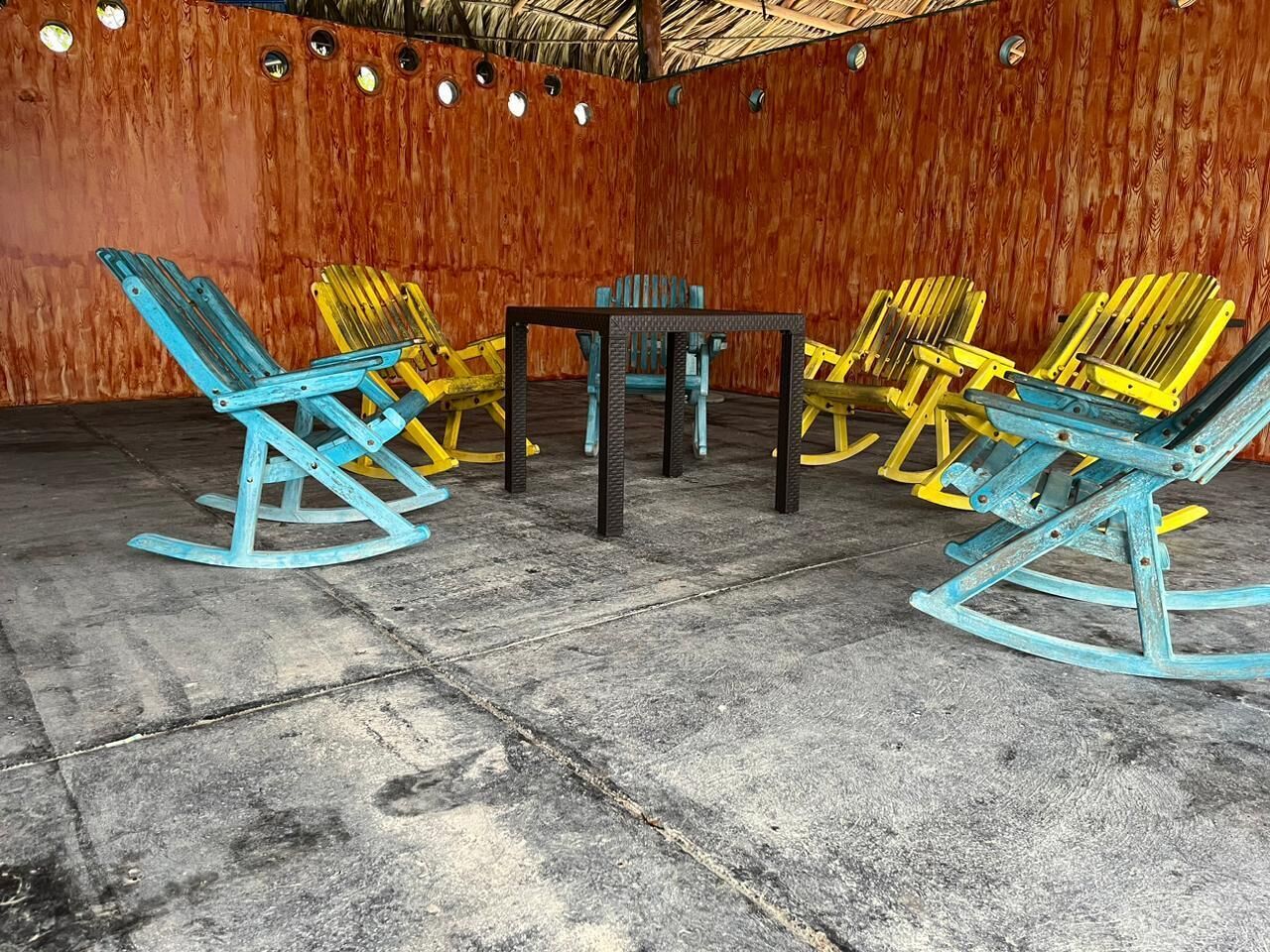 A room with a wooden wall, a concrete floor, and a table surrounded by rocking chairs.