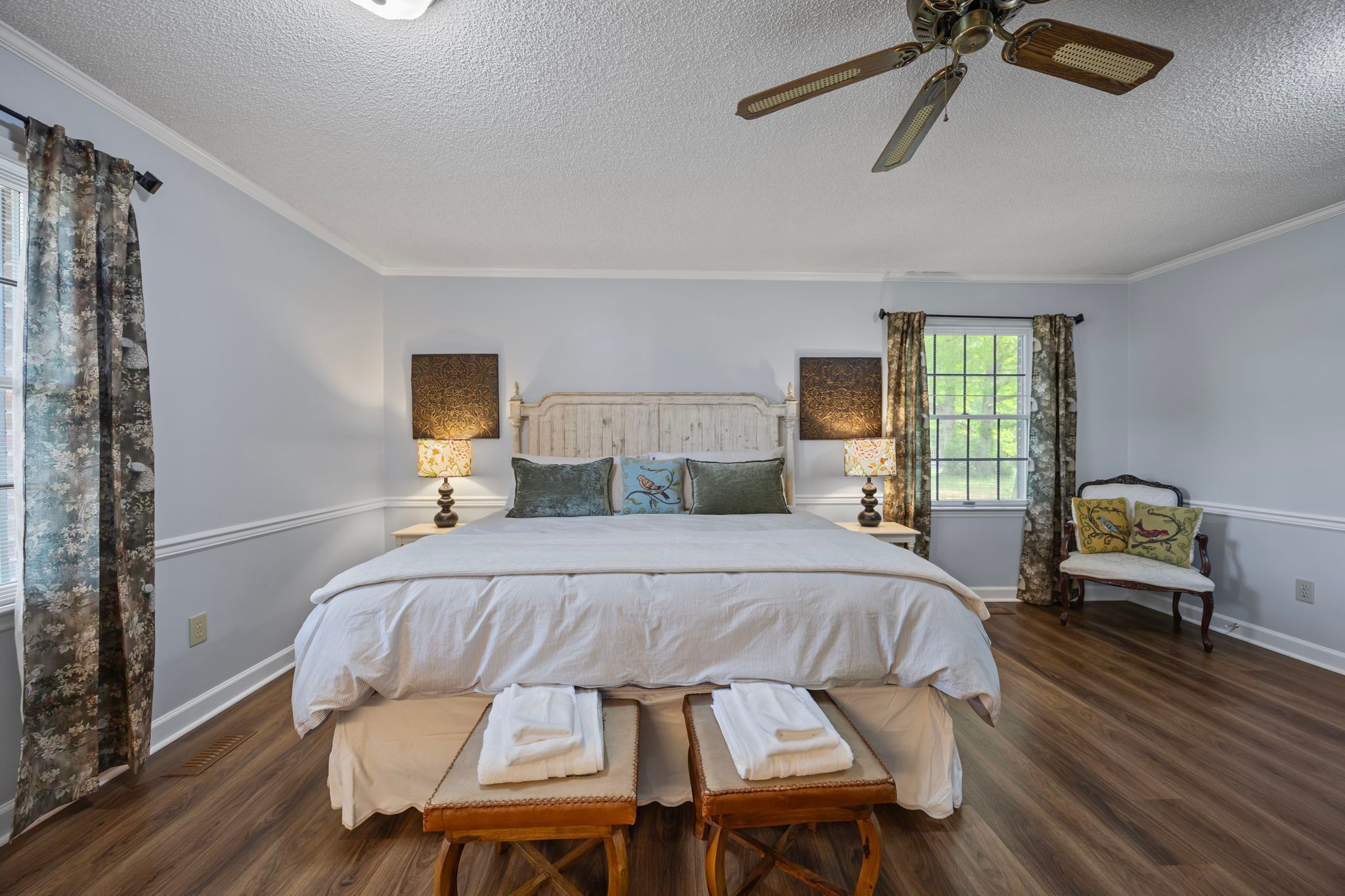 A bedroom with a large bed, two bedside tables, a chair, a ceiling fan, and a window with curtains.