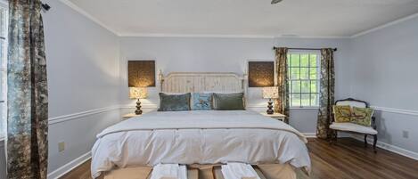 A bedroom with a large bed, two bedside tables, a chair, a ceiling fan, and a window with curtains.