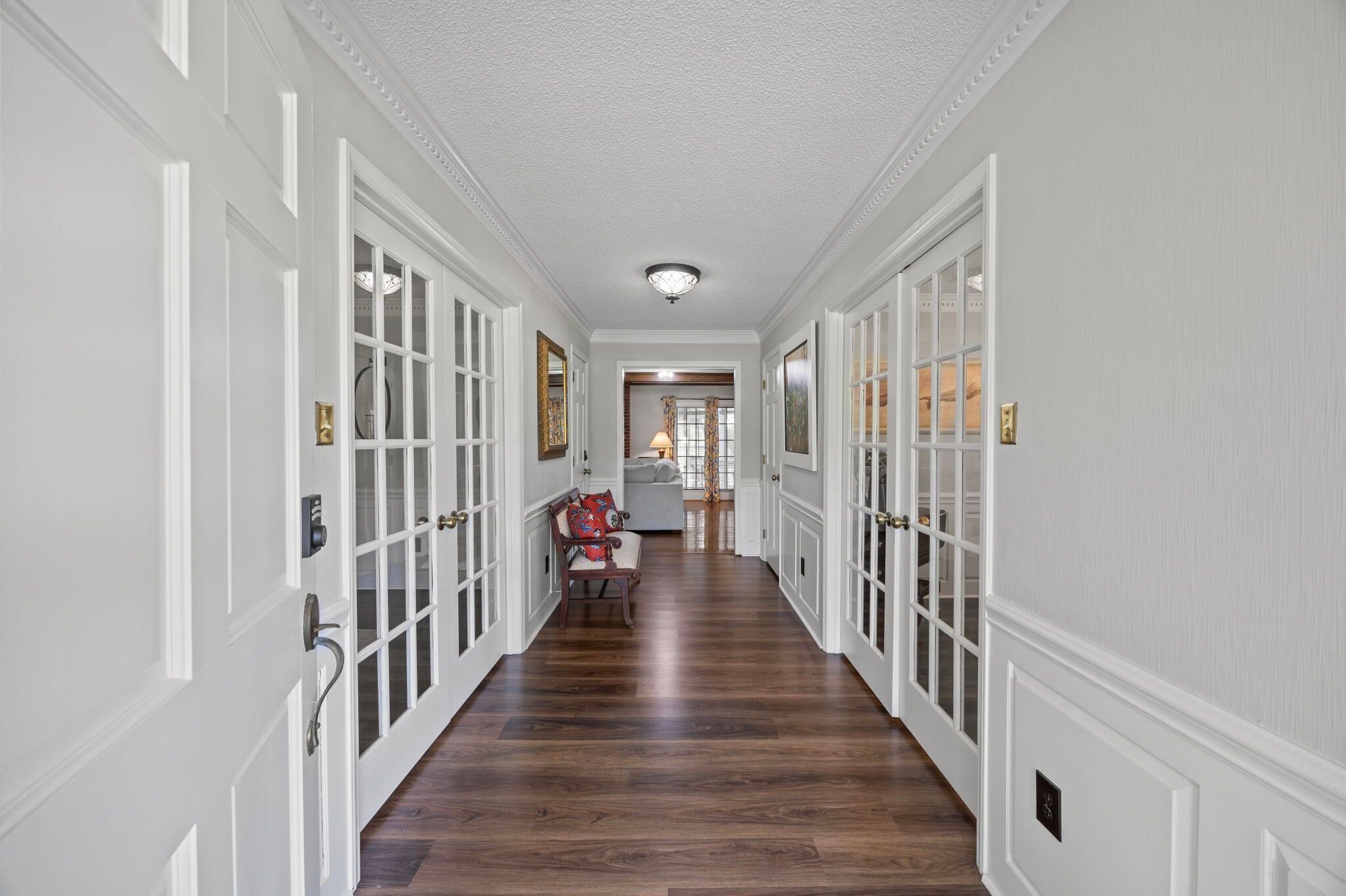 Un pasillo con paredes blancas con paneles y piso de madera, que conduce a una habitación con ventana y puerta.
