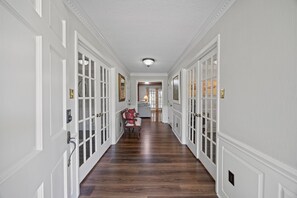 A hallway with white paneled walls and wooden flooring, leading to a room with a window and a door.